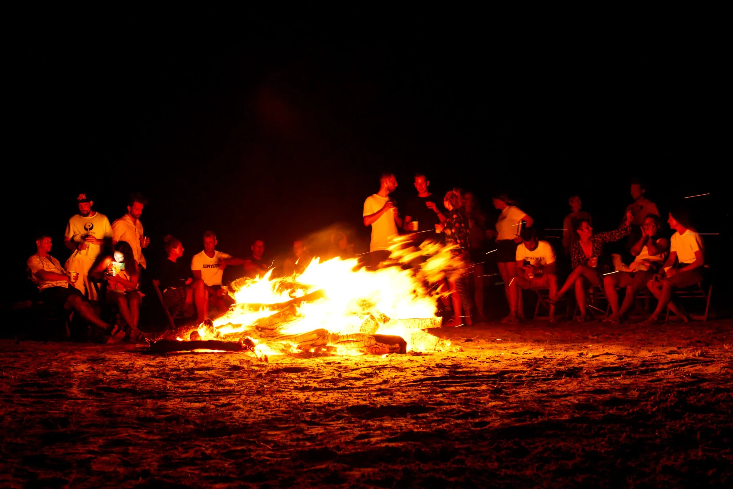Een groep mensen rond een kampvuur op het strand.
