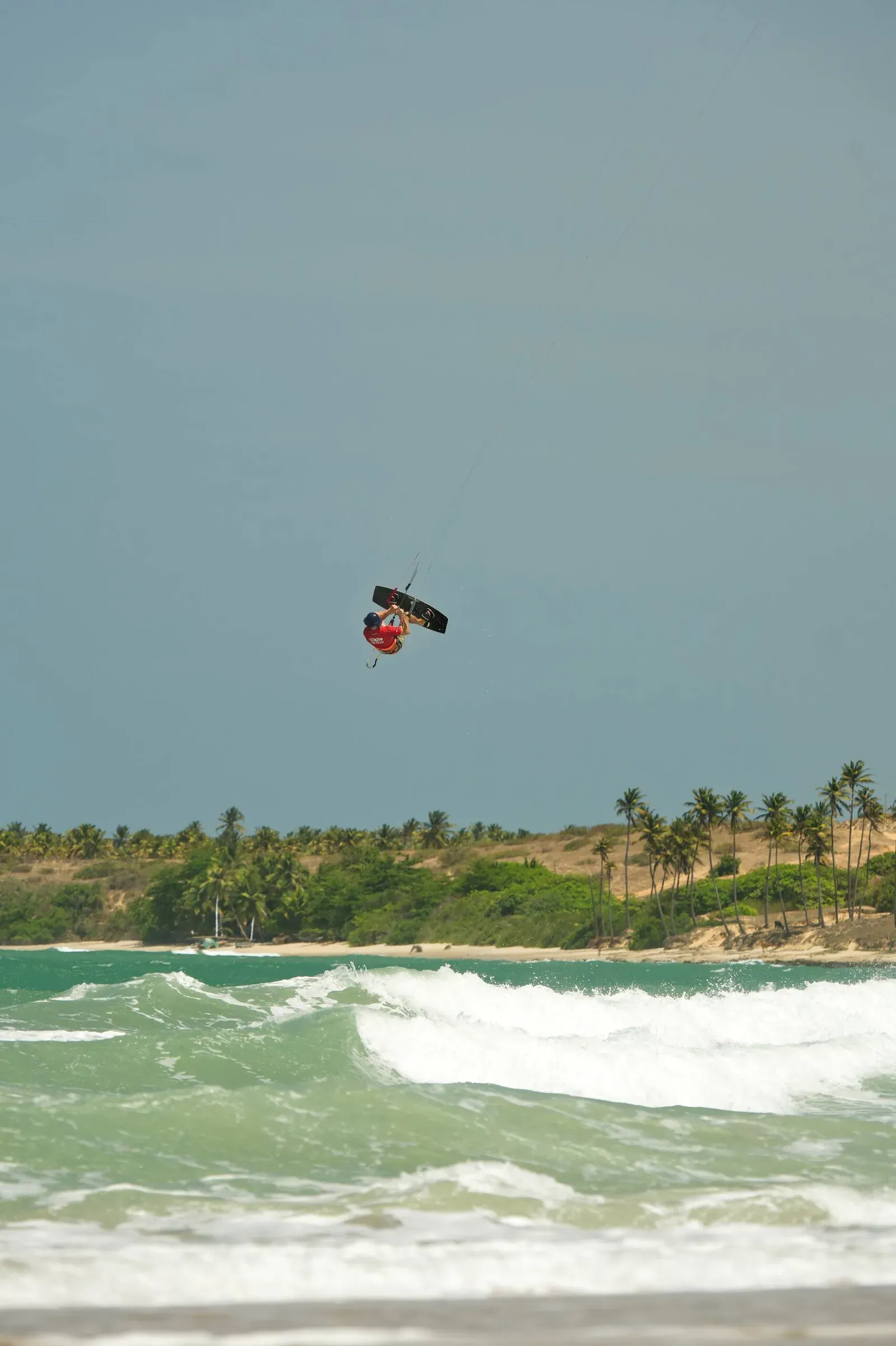 Kitesurfer maakt een hoge sprong boven de golven bij een strand met palmbomen.